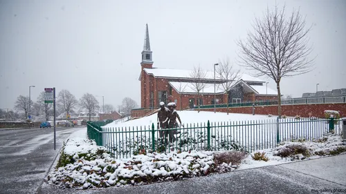 The image shows a morning winter scene featuring the architecture of the Church of Jesus Christ of the Latter Day Saints in Chesterfield, Derbyshire, United Kingdom. Snow is falling gently and covers the church grounds, nearby trees, and a street that runs alongside the property. In the foreground, a statue is positioned behind a green metal fence, partially covered with snow. This is an urban landscape photograph that prominently displays the church, the snow-laden street, and surrounding trees, capturing the quiet atmosphere of Chesterfield during winter.